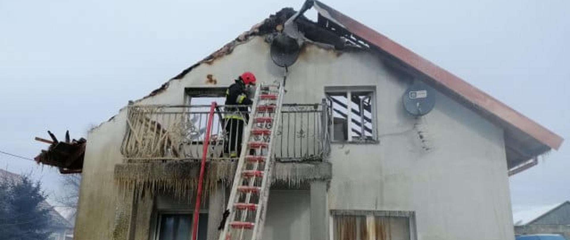 Zdjęcie przedstawia strażaka, w końcowej fazie działań ,znajdującego się na balkonie domu, w którym wybuchł pożar. Budynek ukazany od szczytowej ściany. Zauważyć można zniszczony przez ogień dach. Do balkonu przystawiona jest drabina strażacka a obok niej znajduję się zwisający odcinek węża tłocznego. 