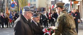 Dwóch mężczyzn w mundurach wyjściowych ochotniczych straży pożarnej w tym Prezes Zarządu Oddziału Powiatowego Związku OSP RP dh Henryk Czubaj oraz Skarbnik dh Ryszard Zyzik składają wiązankę z biało-czerwonymi szarfami na ręce mężczyzny ubranego w mundur wojskowy. W tle uczestnicy uroczystości.