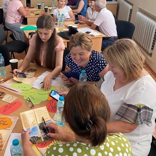 Grupa kilkunastu ludzi siedzi przy kilku stolikach czytając foldery, pisząc flamastrami i uśmiechając się do siebie.