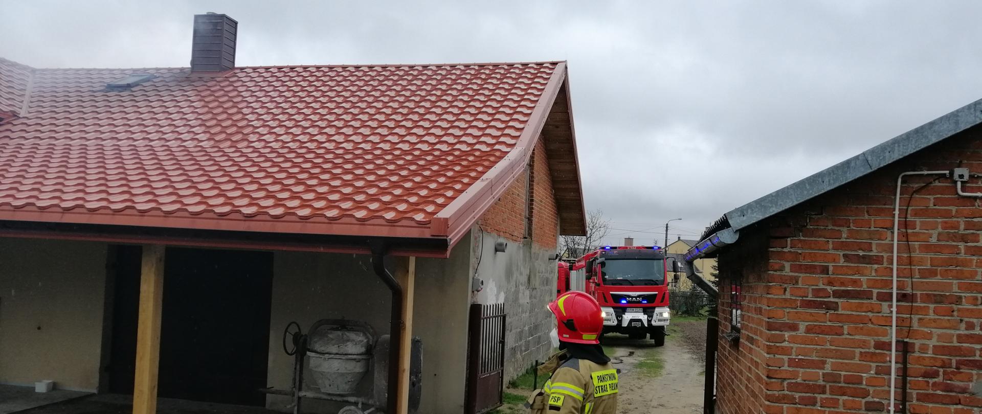 Zdjęcie przedstawia dwa budynki gospodarcze, a między nim znajduje się droga na posesję. Na drodze stoi strażak, w pisakowym stroju strażackim, skierowany bokiem do fotografa, patrząc na komin budynku znajdującego się po lewej stronie. w oddali za strażakiem na drodze widać przód wozu strażackiego.