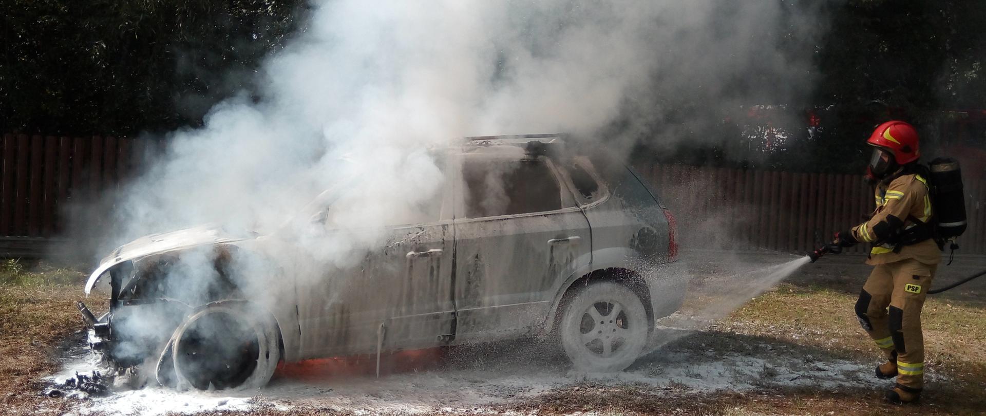 Strażak prowadzący akcję gaszenia pożaru samochodu osobowego. Palący się samochód pokryty jest pianą gaśniczą. Pod podwoziem pojazdu widoczne płomienie. Nad pojazdem unosi się dym.