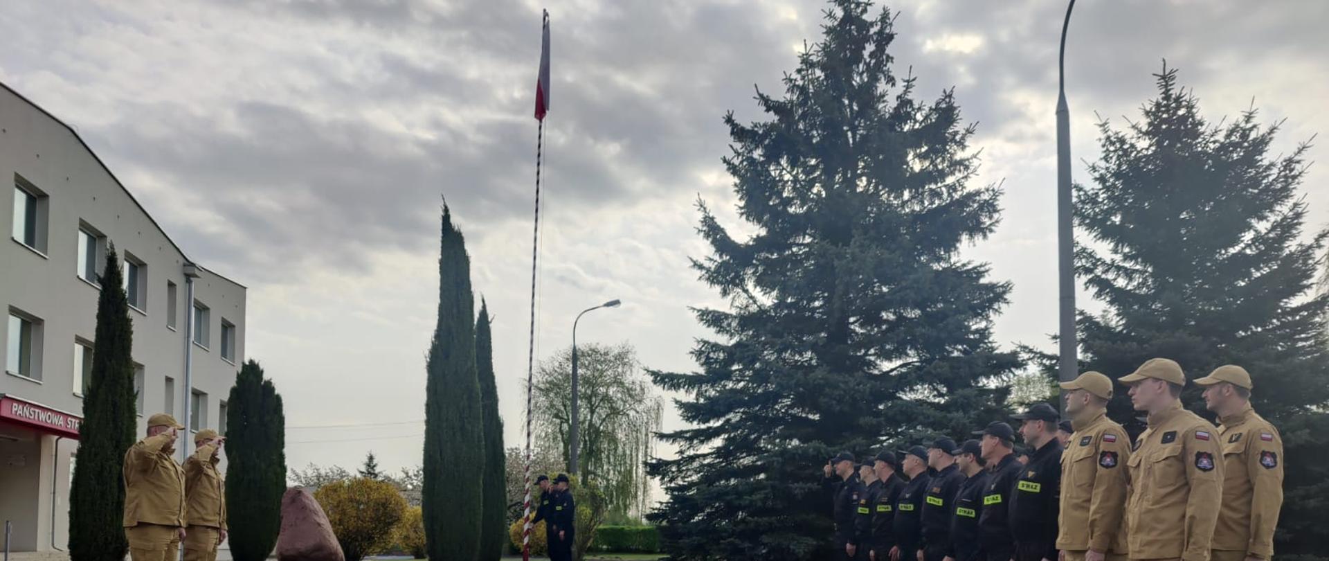 Uroczysta zbiórka przed budynkiem Komendy Powiatowej PSP w Opocznie. Umundurowani strażacy oddają honory w kierunku flagi podniesionej na maszcie. Z lewej strony Komendanci, z prawej kilkunastu funkcjonariuszy, w centralnej części przy maszcie poczet flagowy. 