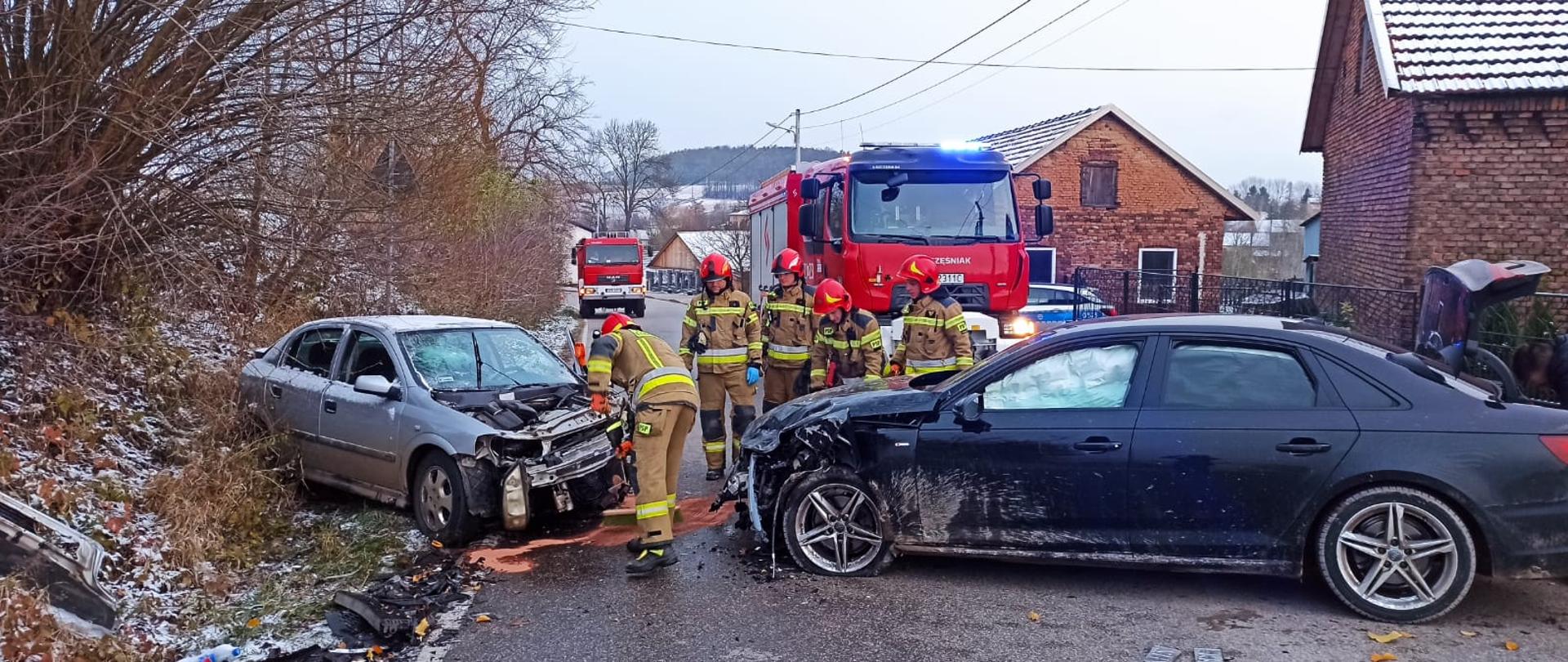 Dwa auta po wypadku znajdują się na jezdni, w tle wozy strażackie oraz pracujący strażacy