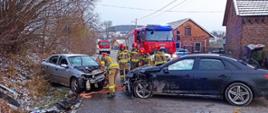 Dwa auta po wypadku znajdują się na jezdni, w tle wozy strażackie oraz pracujący strażacy