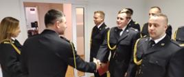Zdjęcie przedstawia funkcjonariuszy podczas uroczystości mianowania na wyższe stanowiska służbowe. Strażacy stojący w dwuszeregu, znajdują się w świetlicy Komendy Powiatowej Państwowej Straży Pożarnej w Lwówku Śląskim. Komendant Powiatowy PSP w Lwówku Śląskim wręcza mianowanie funkcjonariuszowi. 