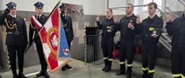 Zdjęcie przedstawia 3 strażaków wchodzących w skład pocztu sztandarowego oraz 4 strażaków, którzy składają ślubowanie. Jeden ze strażaków z pocztu trzyma w ręce sztandar Komendy Powiatowej PSP w Gostyniu. Strażacy z pocztu ubrani są w umundurowanie reprezentacyjne, a pozostali strażacy w koszarowe. Strażacy w pocztu oddają honor poprzez salutowanie.