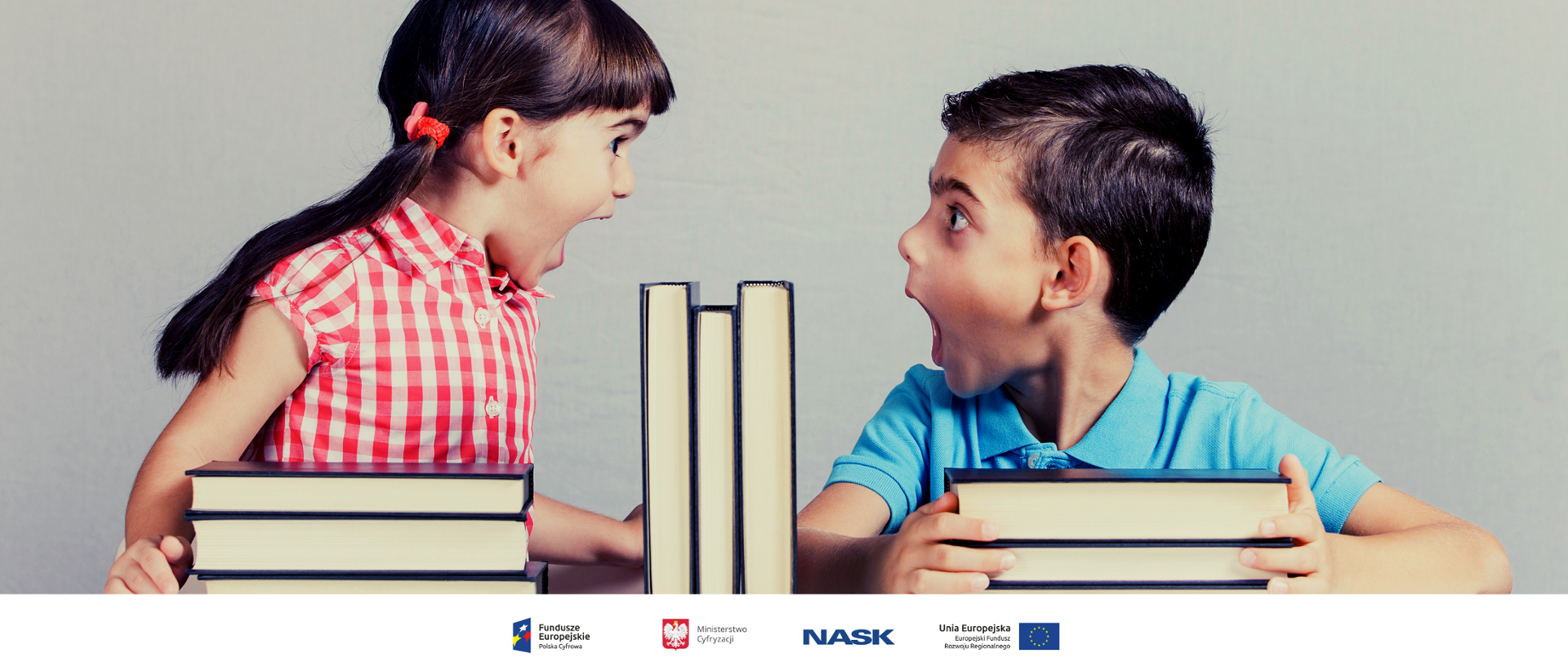 Kolorowo ubrani dziewczynka i chłopiec stroją miny patrząc na siebie. Na stoliku przed nimi leży kilka książek