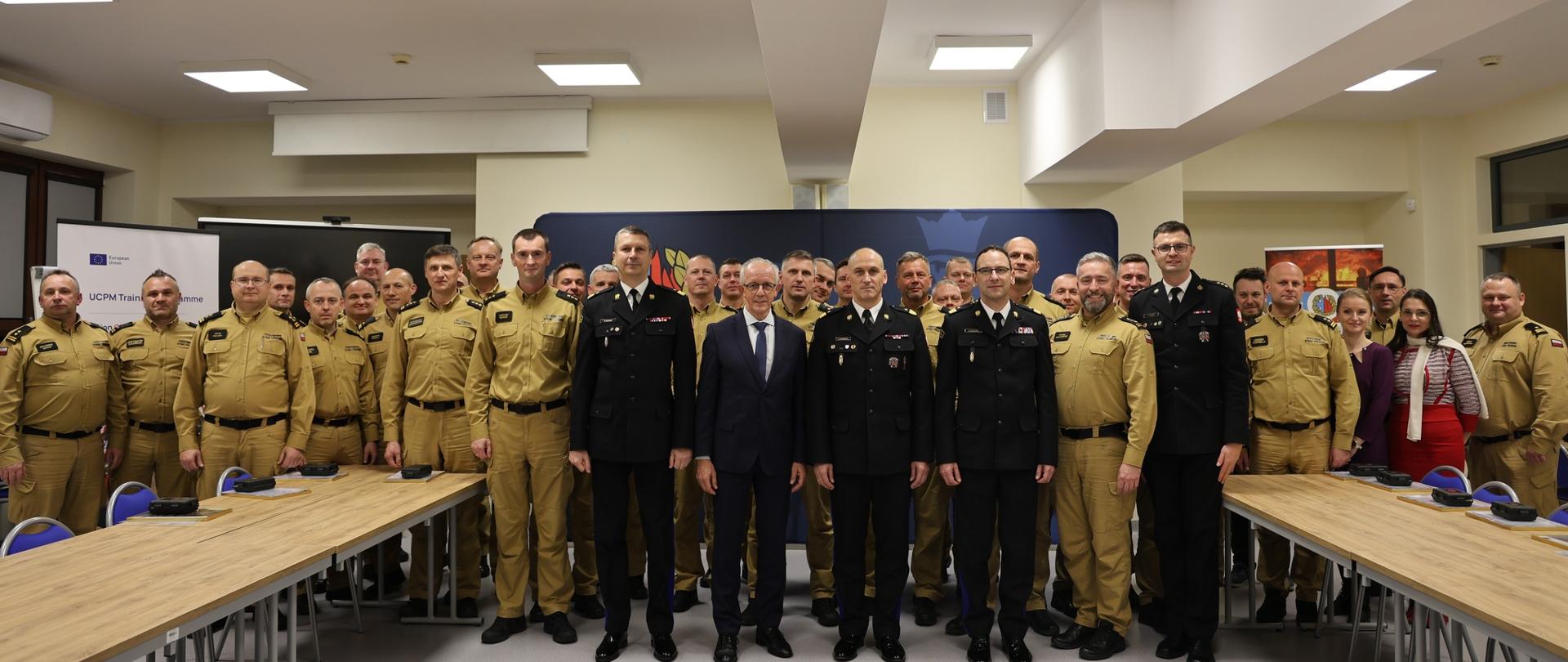 Duża grupa osób w sali – na przedzie kadra w galowych mundurach PSP i mężczyzna w garniturze, w tle uczestnicy w mundurach służbowych koloru żółtego, głównie strażacy PSP.
