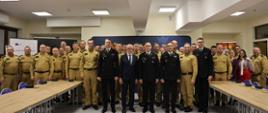 Duża grupa osób w sali – na przedzie kadra w galowych mundurach PSP i mężczyzna w garniturze, w tle uczestnicy w mundurach służbowych koloru żółtego, głównie strażacy PSP.
