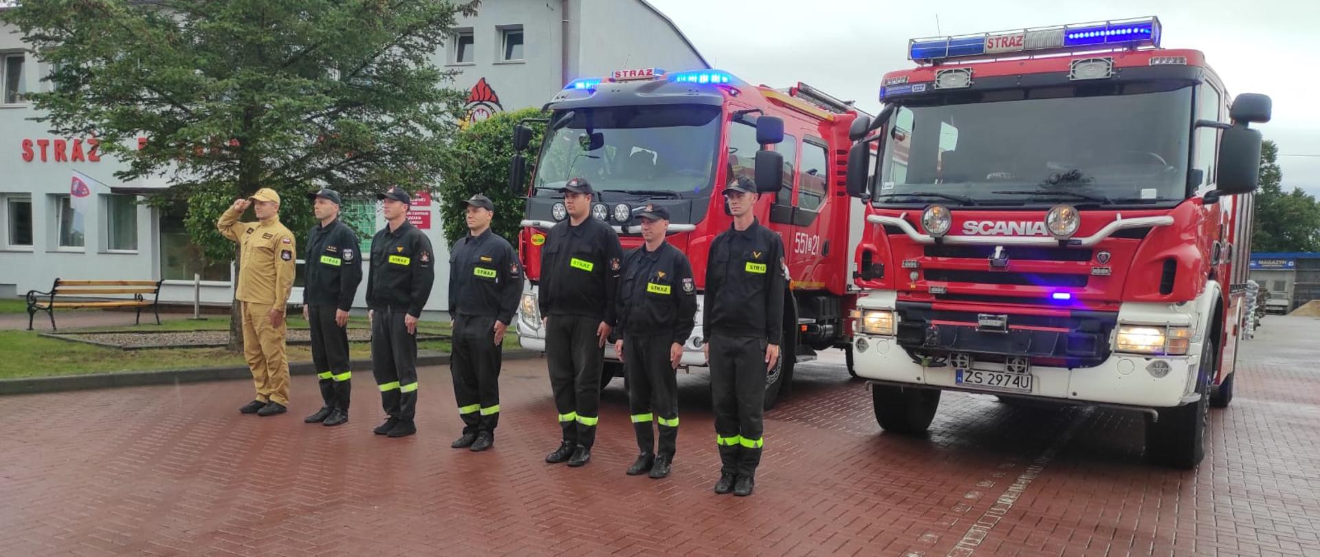 Zdjęcie przedstawia dwa wozy strażackie przed remizą psp oraz strażaków w szeregu podczas Hołd Powstańcom w 79. rocznicę wybuchu Powstania Warszawskiego