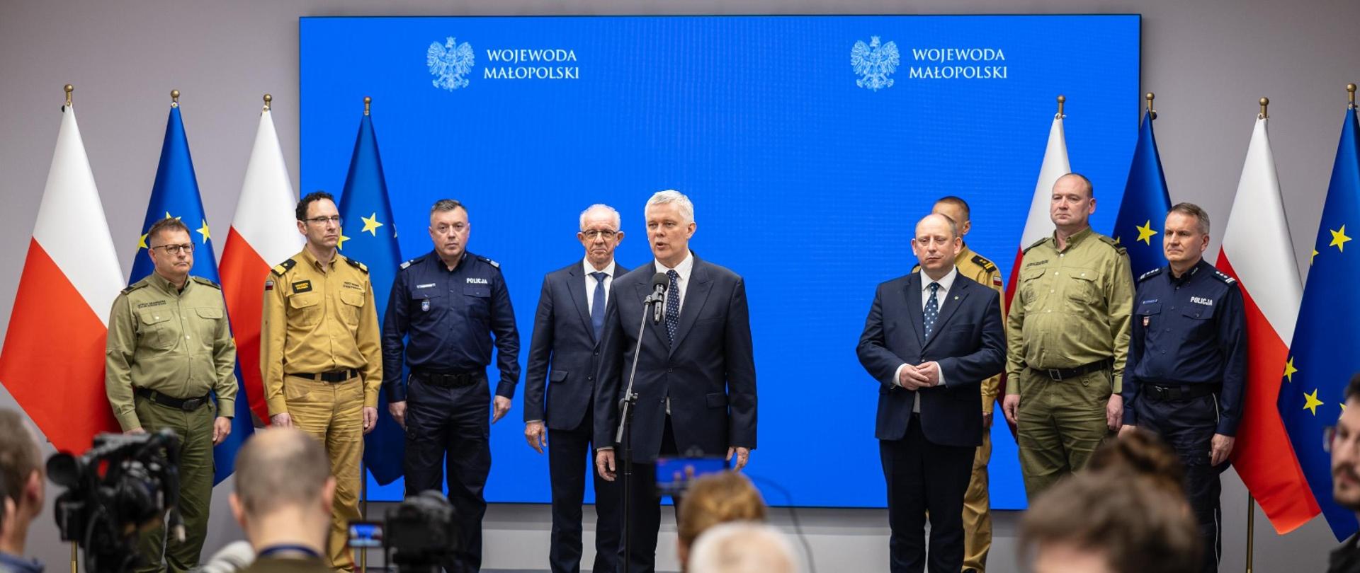 Na zdjęciu widoczna jest konferencja prasowa z udziałem wysokich rangą przedstawicieli służb mundurowych oraz administracji państwowej. Na środku, przed mikrofonem, przemawia mężczyzna w garniturze i białej koszuli. Za nim stoi kilku urzędników oraz funkcjonariuszy w mundurach straży pożarnej, policji i straży granicznej. W tle znajduje się niebieski ekran z napisem "Wojewoda Małopolski" oraz flagi Polski i Unii Europejskiej. Na pierwszym planie widoczni są dziennikarze i fotoreporterzy rejestrujący wydarzenie.