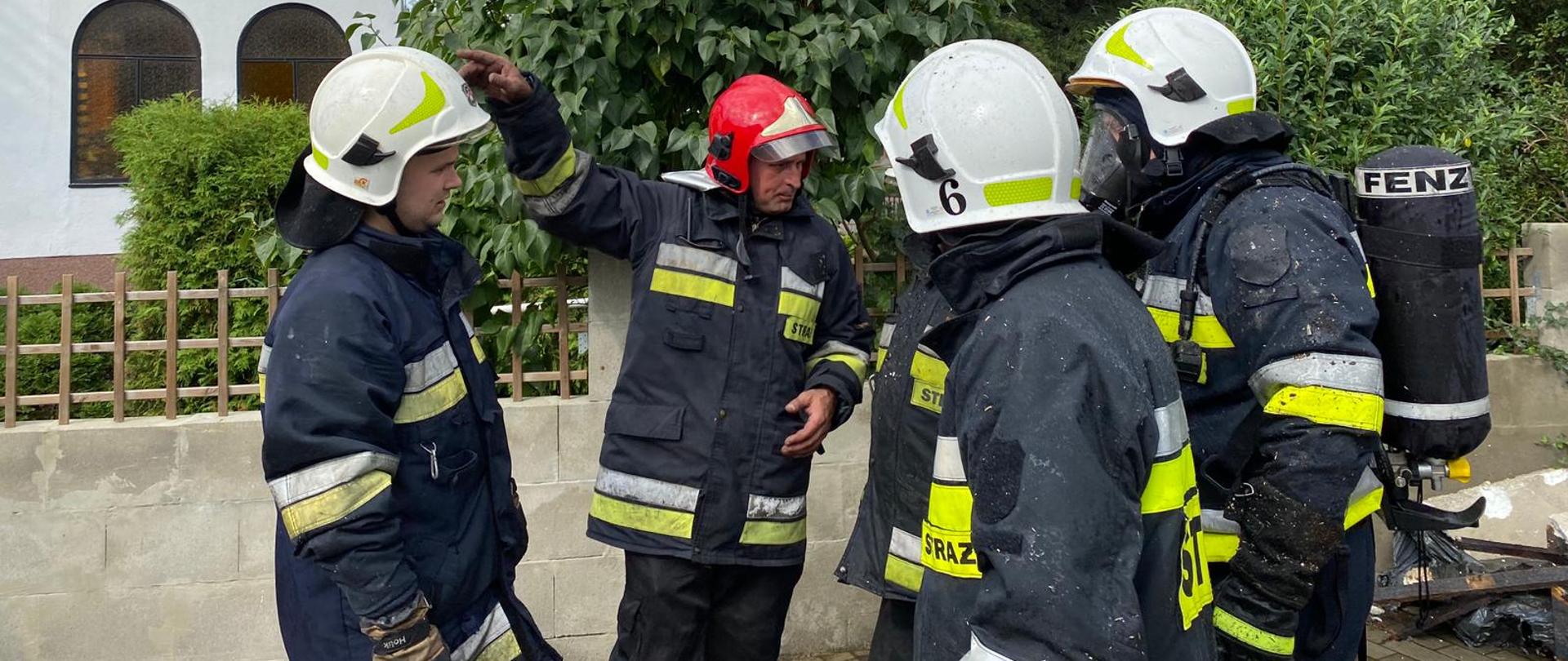 4 strażaków w ubraniach specjalnych koloru czarnego z białymi i żółtymi elementami przygotowują się do działań