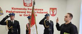 Na zdjęciu w mundurach galowych po lewej stronie poczet sztandarowy ze sztandarem. Po prawej strażak składający ślubowanie w ubraniu czarnym koszarowym. Uroczystość ślubowania ma miejsce w sali konferencyjnej.