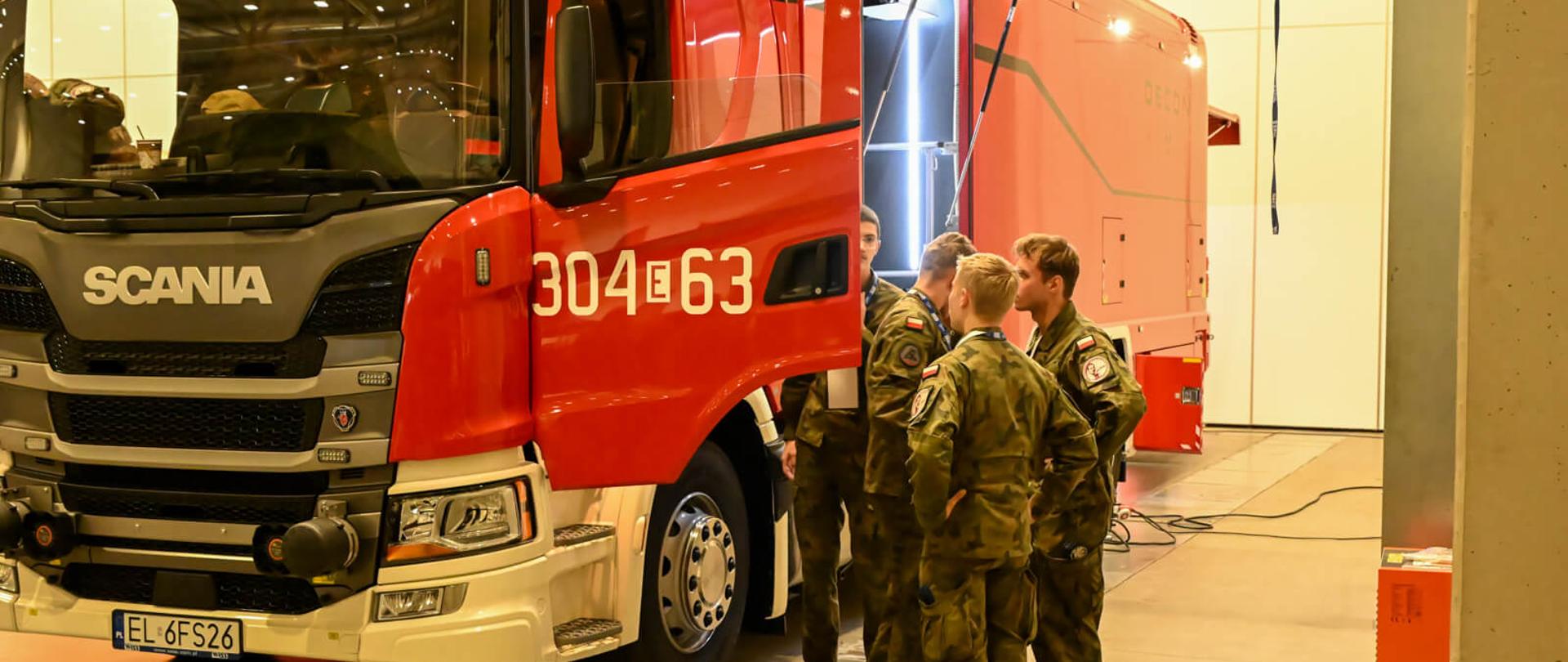 Młodzież w umundurowaniu wojskowym ogląda ciężki samochód specjalny Scania należący do Państwowej Straży Pożarnej, prezentowany podczas targów. Uczestnicy zapoznają się z wyposażeniem pojazdu i jego zabudową specjalistyczną.
