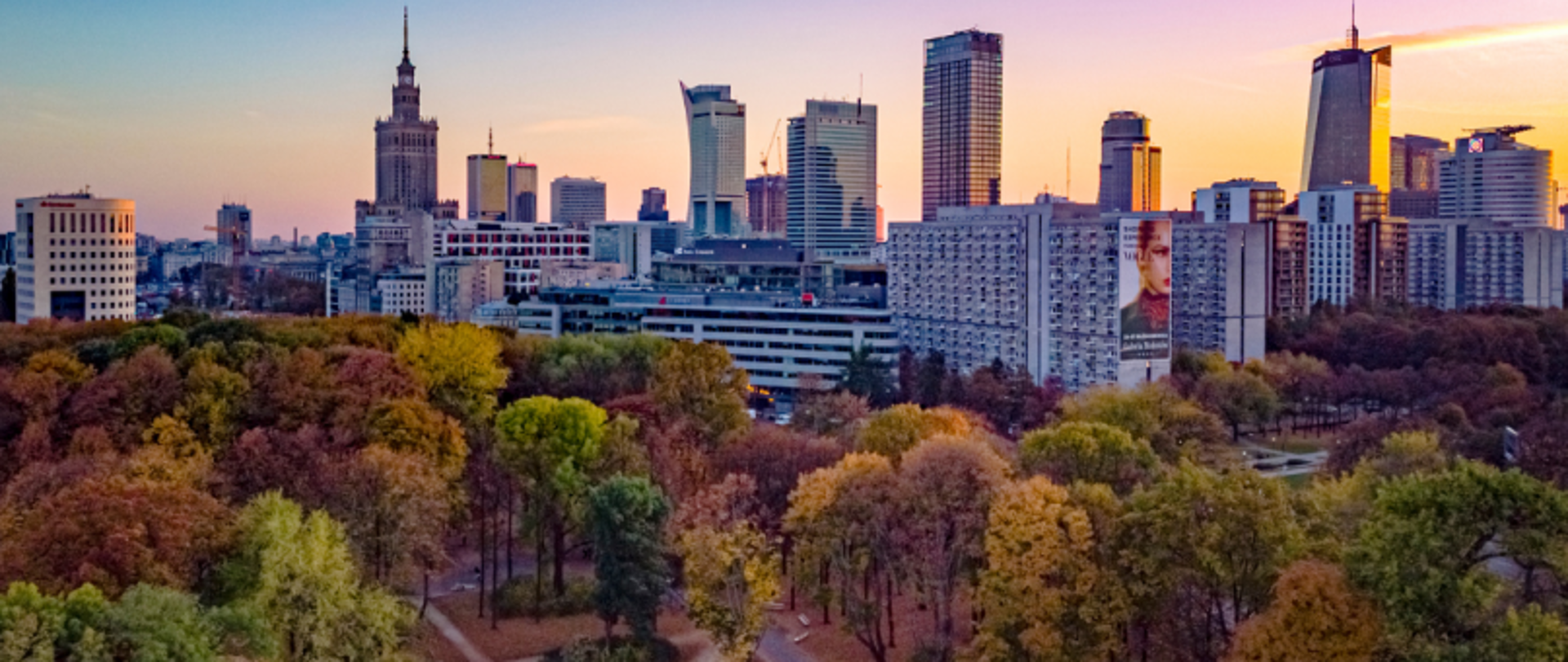 Cityscape of Warsaw at sunset with the Palace of Culture and Science and surrounding buildings; autumn-colored park in the foreground and orange-purple sky in the background.