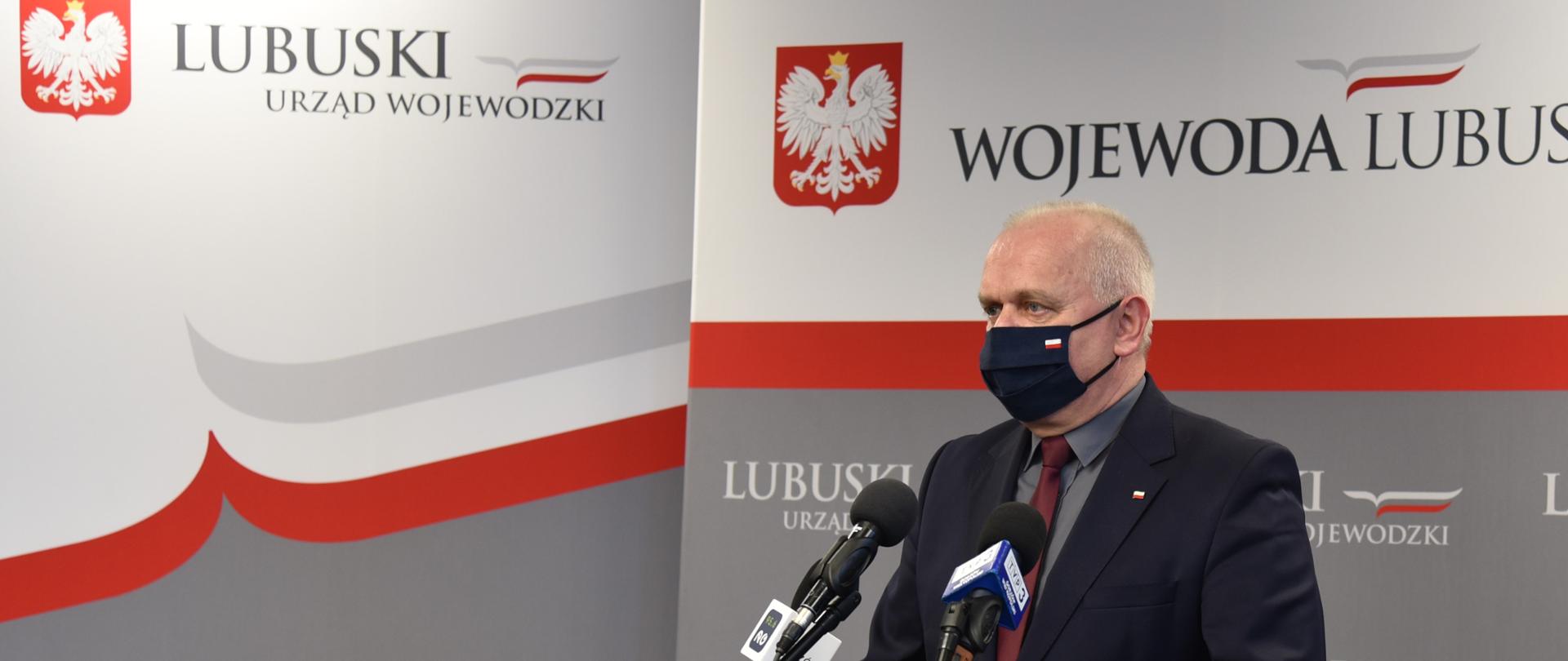 Wojewoda Władysław Dajczak stoi w ciemnym garniturze przed ustawionymi na stojakach mikrofonami. W tle, za nim stoją ścianki promocyjne urzędu i wojewody utrzymane w biało-czerwono-szarej kolorystyce. 