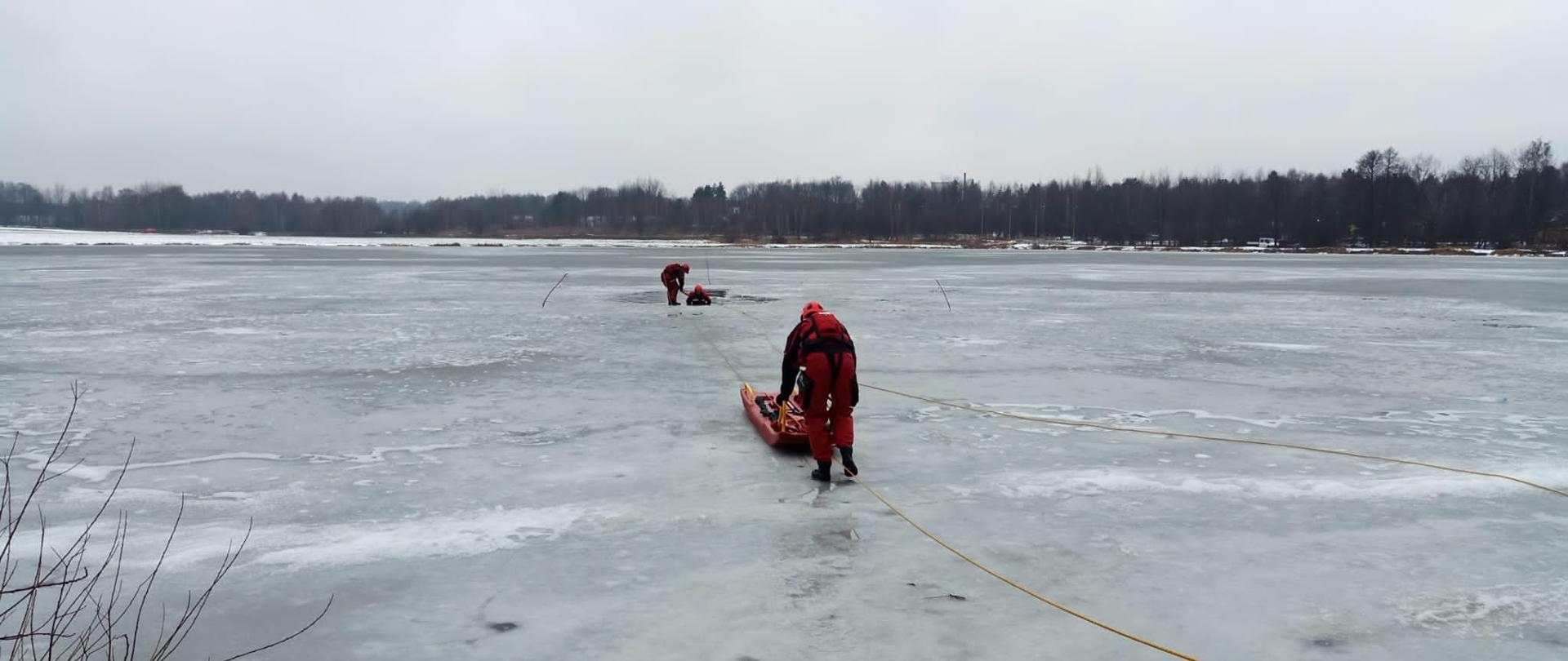 Dwóch strażaków w oddali w czerwonych skafandrach z których jeden jest w wodzie. Trzeci strażak idzie do nich z deską lodową przypięty do liny. 