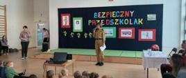 Pani strażak ubrana w mundur służbowy w kolorze musztardowym trzyma w dłoni mikrofon, przez który przemawia do zgromadzonych przed nią dzieci. Za nią znajduje się czarna ścianka, na której znajduje się kolorowy napis "BEZPIECZNY PRZEDSZKOLAK" a dokoła napisu przyczepione są rysunki dzieci o tematyce strażackiej.