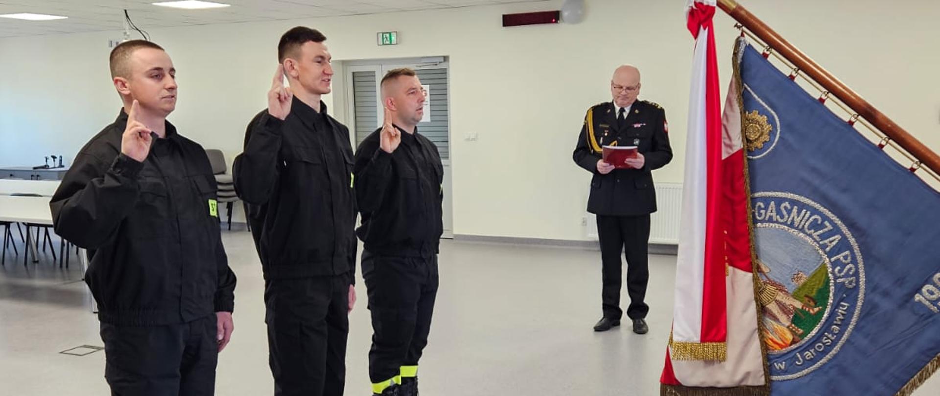 Trzech młodych strażaków w czarnych mundurach składa uroczystą przysięgę na sztandar straży pożarnej. Przed nimi stoi funkcjonariusz w galowym mundurze, trzymający sztandar. W tle widoczny jest oficer odczytujący tekst przysięgi.