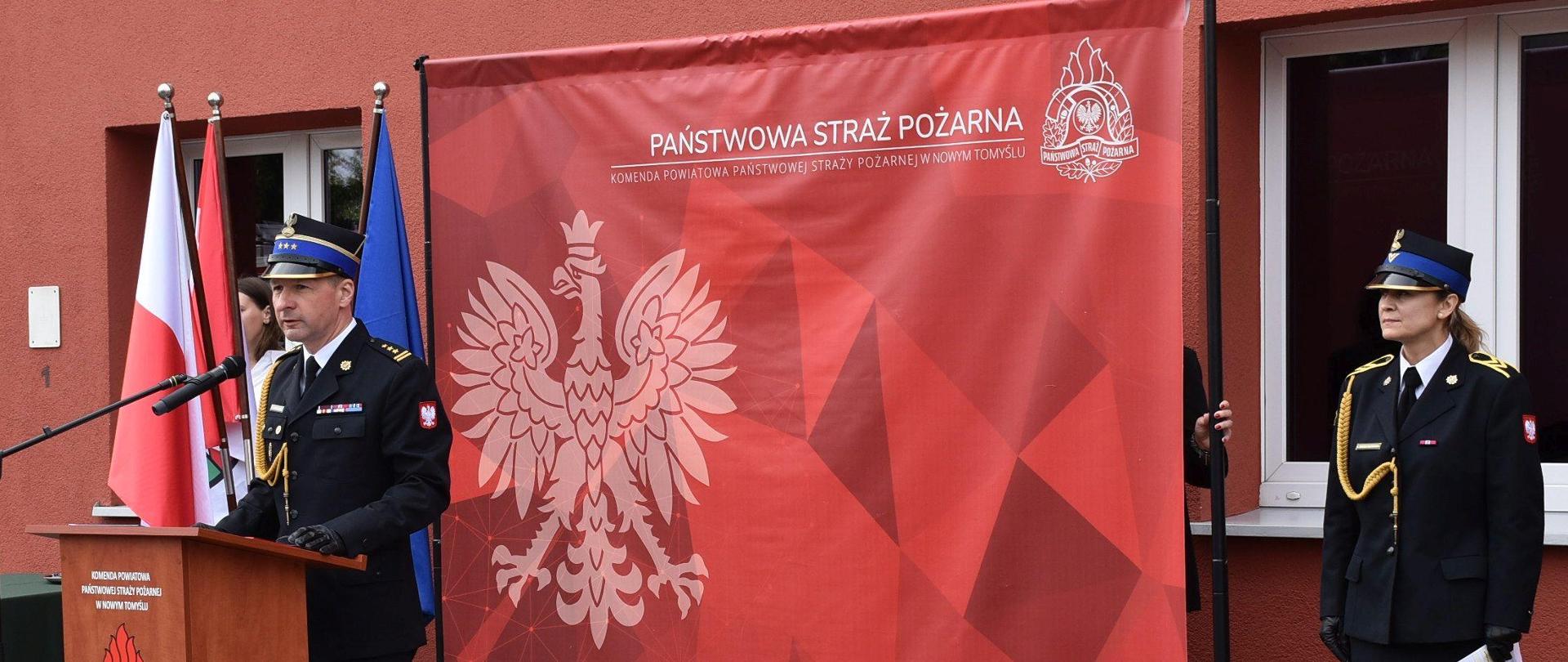 przemówienie Zastępcy Wielkopolskiego Komendanta Wojewódzkiego Państwowej Straży Pożarnej, w tle duża czerwona ścianka z orłem, obok stoi kobieta strażak
