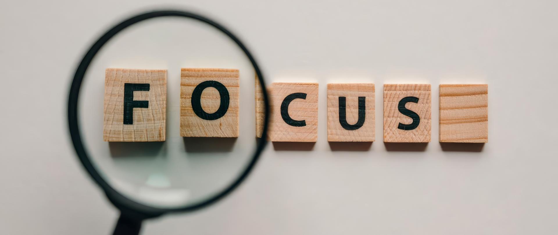 Na szarym tle leżą drewniane klocki z literami układające się w napis FOCUS . Pierwsze dwie litery powiększone przez lupę.