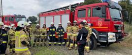 strażacy ubrani w ubrania specjalne stoją na tle samochodu pożarniczego wsłuchują się w słowa strażaka w czarnym ubraniu