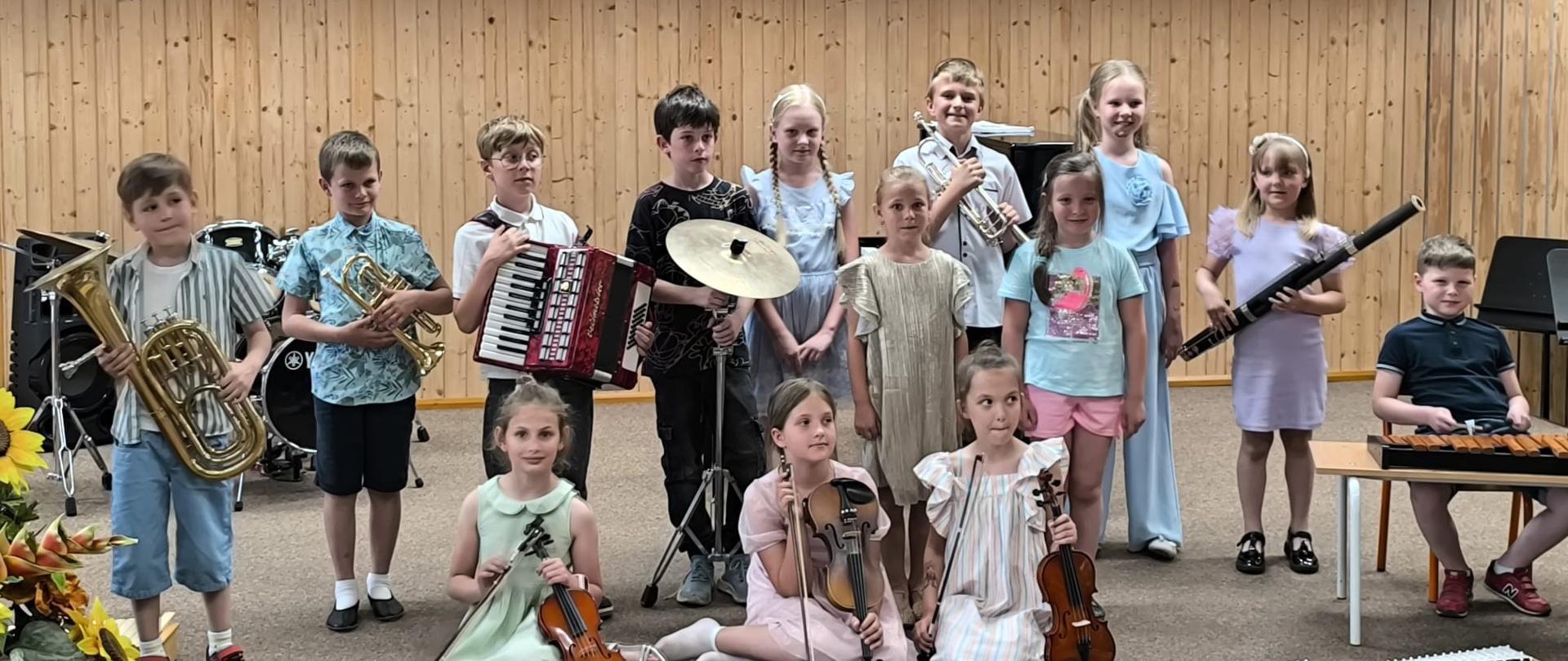 Kilkanaścioro dzieci stoi lub siedzi na scenie ze swoimi instrumentami.