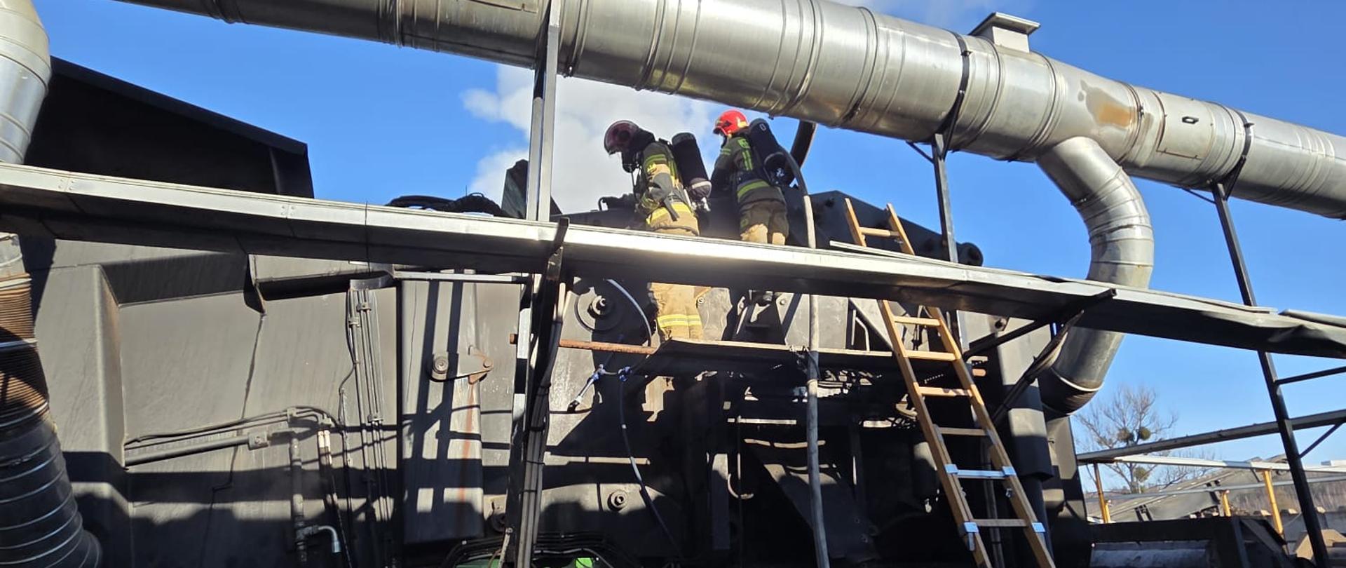 Dwóch strażaków w odzieży ochronnej stoi na metalowej platformie przy dużej instalacji przemysłowej z rurami wentylacyjnymi; obok oparta drabina, a w tle widać unoszącą się parę lub dym na tle bezchmurnego, niebieskiego nieba.