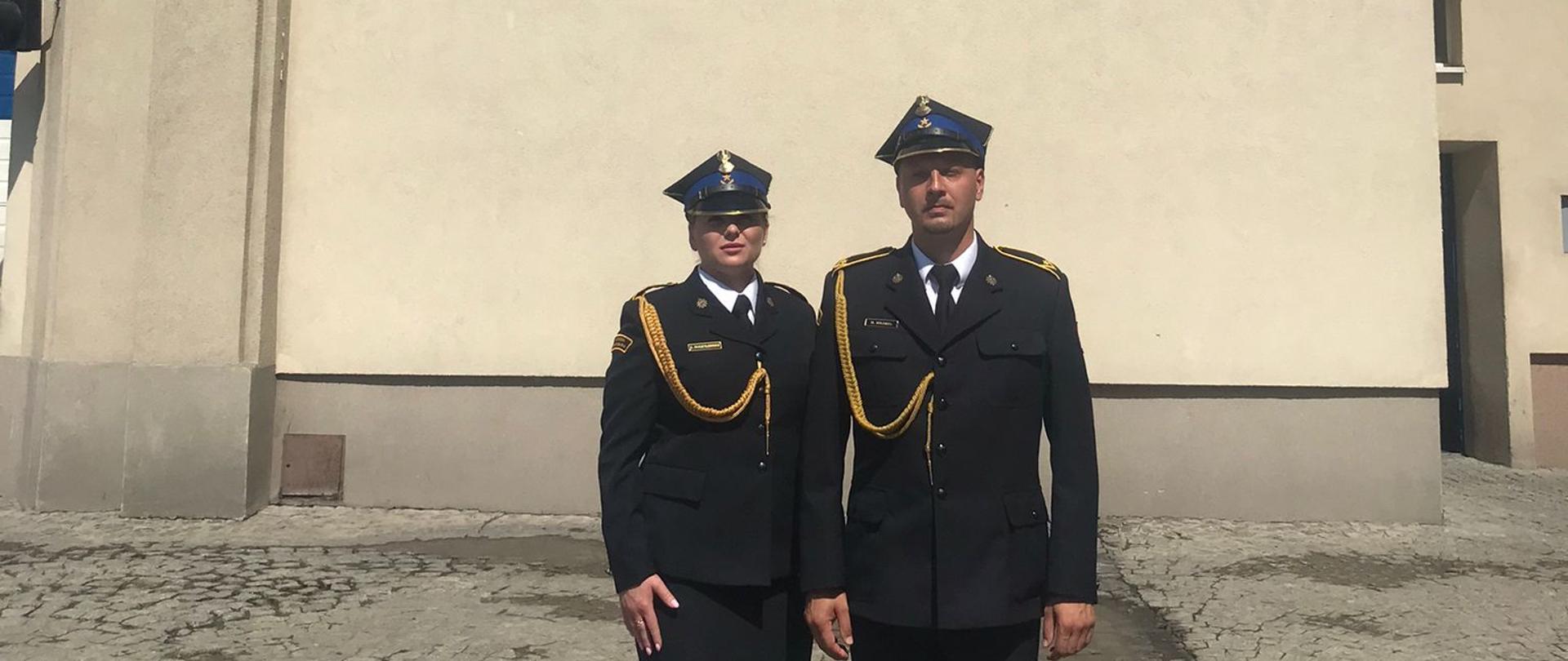 Dwoje funkcjonariuszy ubranych w czarny mundur galowy ze złotym sznurem i rogatywką. Od lewej funkcjonariuszka, po prawej funkcjonariusz. Zdjęcie zrobione na tle budynku szkoły z 3 różnymi odznaczeniami szkoły. Plac wyłożony szarą kostką. 