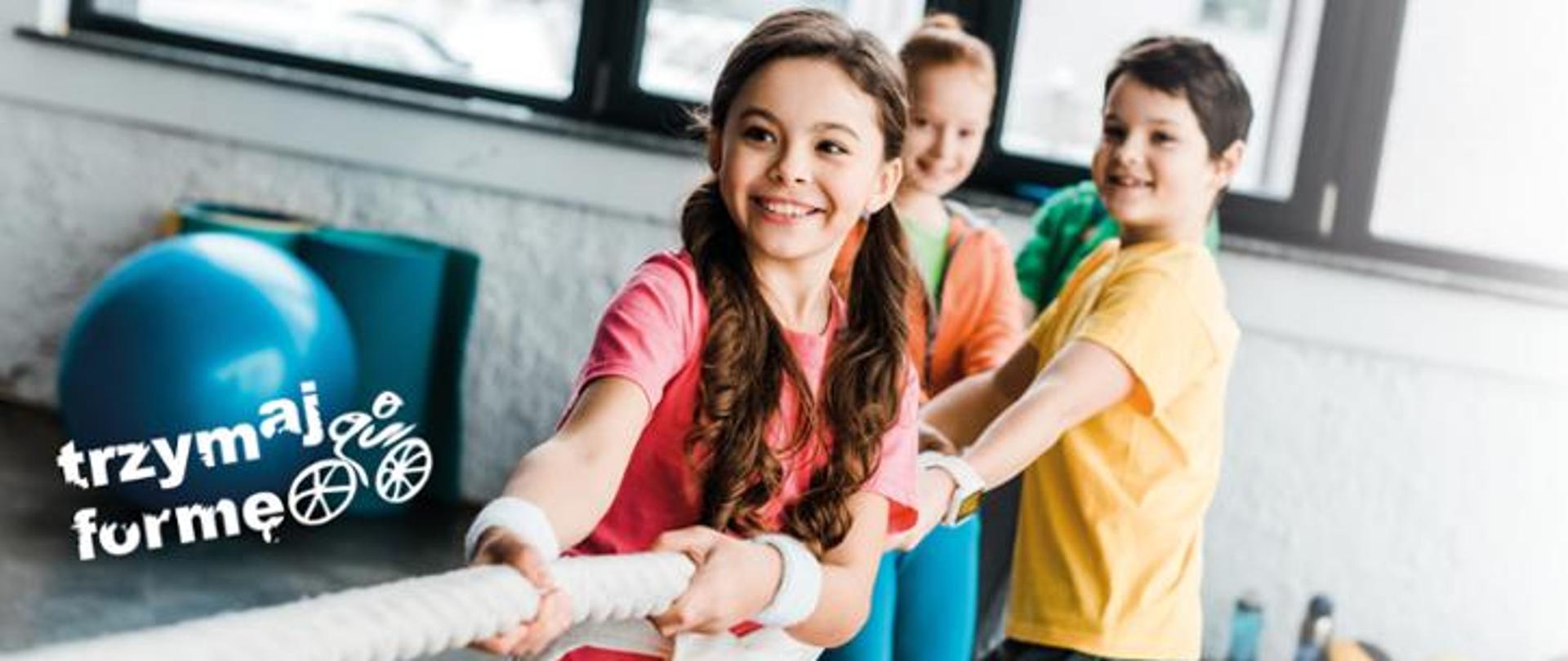 zdjęcie przedstawia troje dzieci . Na pierwszym planie dziewczynka kolejno chłopiec i kolejno dziewczynka. Wszyscy trzymają linę, którą przeciągają w swoją stronę. W tle duża niebieska piłka do ćwiczeń i napis: trzymaj formę