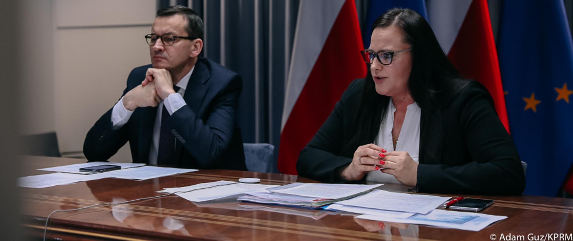 Premier Mateusz Morawiecki oraz minister Małgorzata Jarosińska-Jedynak siedzą przy stole konferencyjnym, w tle flagi UE i Polski
