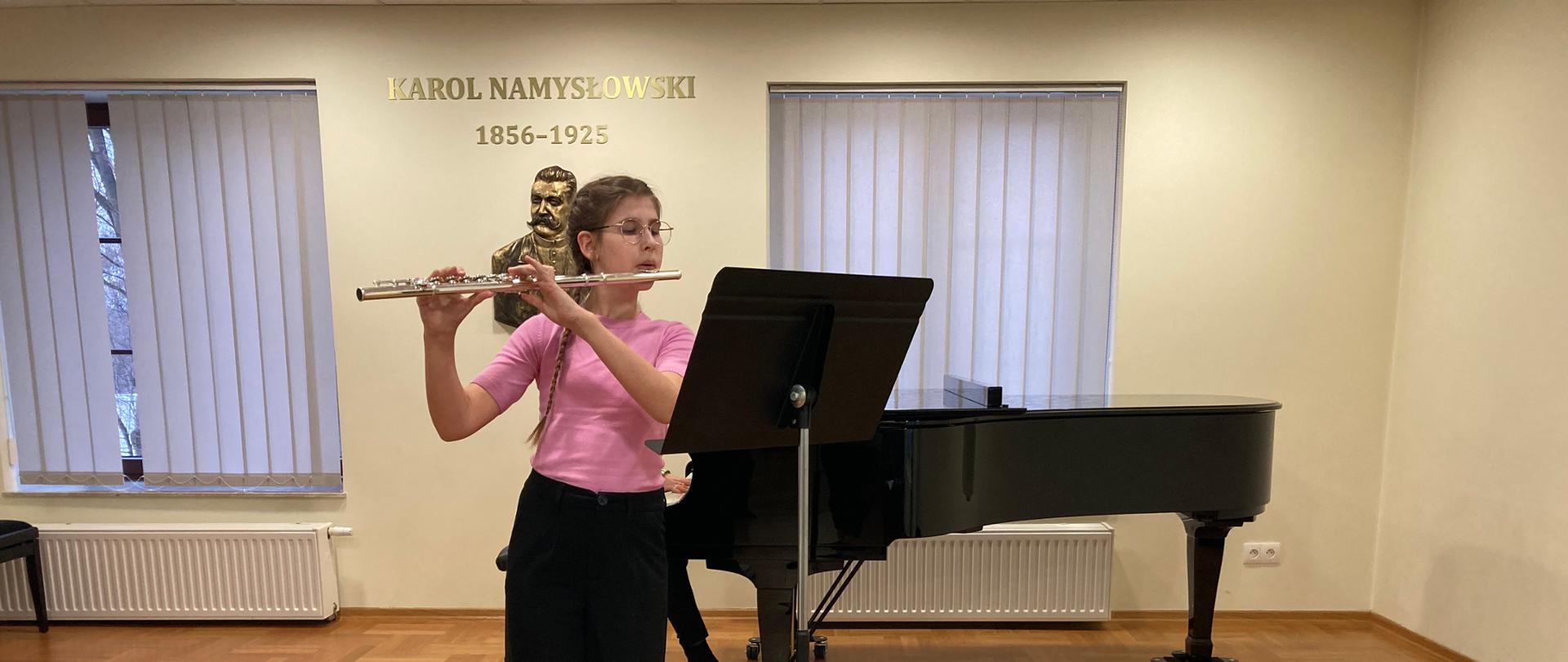 Popis uczniów klasy IV cyklu 6-letniego w dniu 21 marca 2023 r. w sali koncertowej PSM I st. im. K. Namysłowskiego w Szczebrzeszynie. Na scenie uczennica klasy fletu Kornelia Duda, nauczyciel Katarzyna Kuchta. Gra na flecie, stoi na scenie, przed nią stoi pulpit. Z tyłu za nią stoi czarny fortepian na którym akompaniuje Alicja Słupska. Uczennica ubrana w różową bluzkę i czarne spodnie. Na ścianie napis Karol Namysłowski 1856-1925. 