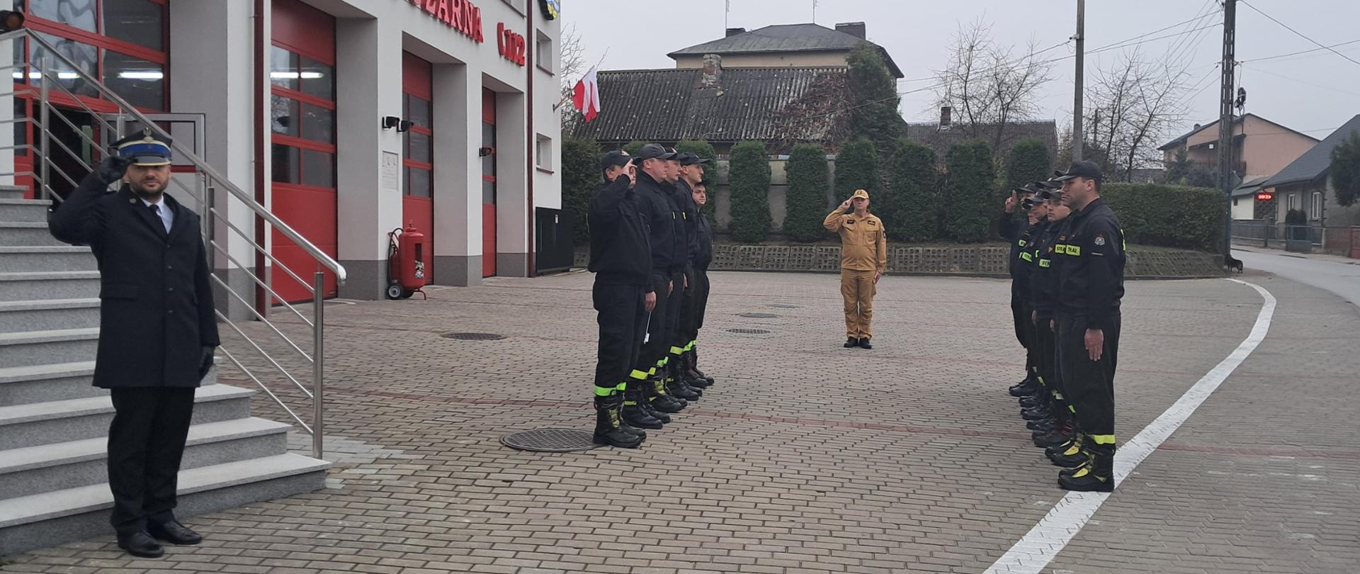 Na zdjęciu widoczna grupa strażaków w umundurowaniu koszarowym i służbowym stoi na placu przed budynkiem Komendy Powiatowej PSP w Przysusze. Po lewej stronie, na dole schodów, stoi mężczyzna w galowym mundurze salutujący. Przed nim stoi szpaler strażaków, a na końcu, na wprost nich, w innej pozycji salutuje strażak w jasnym umundurowaniu. W tle widać budynek komendy z czerwonymi drzwiami garażowymi oraz budynki mieszkalne.