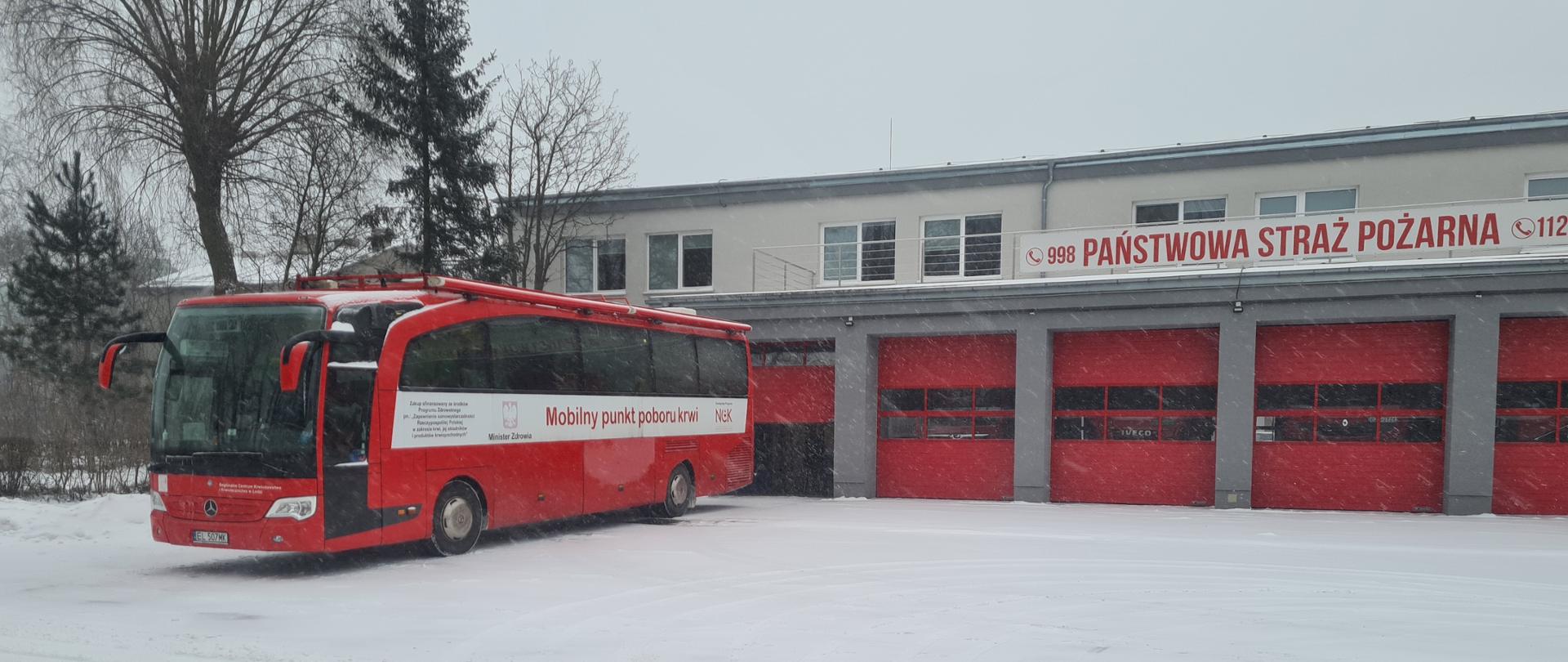 Zdjęcie przedstawia czerwony autokar zwany mobilnym punktem poboru krwi na tle czerwonych bram garażowych Jednostki Ratowniczo - Gaśniczej w Zgierzu