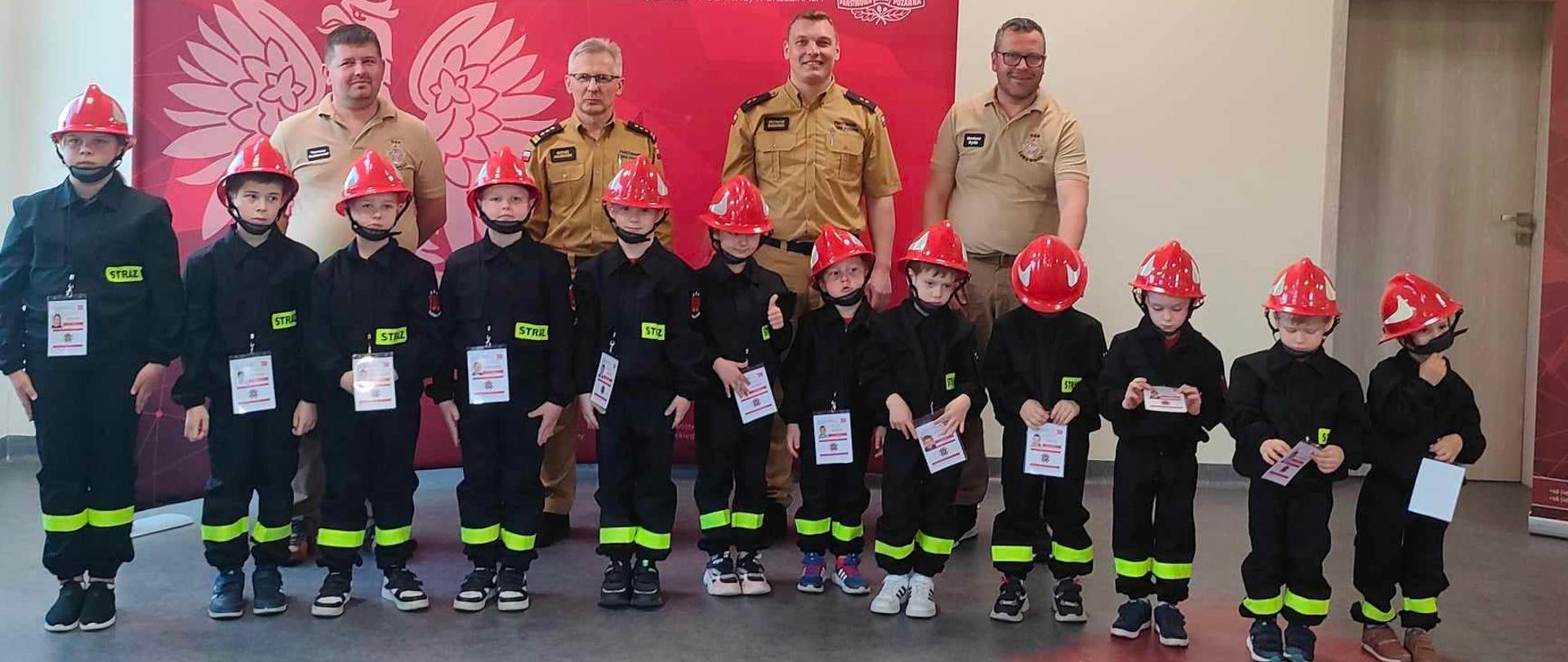 sala konferencyjna komendy powiatowej psp w brzezinach w szeregu stoją po ślubowaniu dzieci z DDP z OSP Lubowidza za nimi komendant powiatowy psp w brzezinach starszy brygadier mariusz kaczmarski , naczeik wydziału operacyjno szkoleniowego Krzysztof Bogdański oraz zarząd OSP Lubowidza