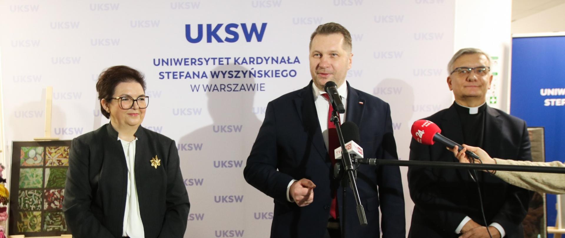 Minister Czarnek stoi pomiędzy ubranymi na czarno kobietą i mężczyzną, mówi do kilku mikrofonów, za nim ścianka z napisem UKSW.