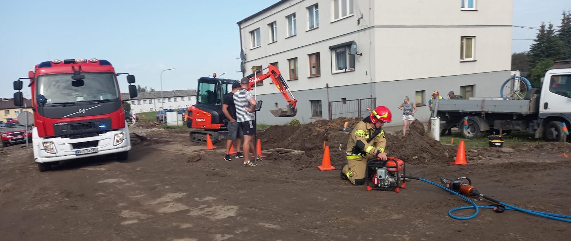 Zdjęcie przedstawia strażaka obsługującego pompę hydrauliczną w tle samochody pożarnicze, koparka i pracownicy geodezyjni.