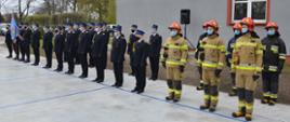 W szyku na baczność jeden obok drugiego stoją strażacy w mundurach wyjściowych kolory czarnego oraz umundurowaniu specjalnym koloru piaskowego i czerwonych hełmach. W tle jeden z budynków Komendy, ściana koloru szarego.