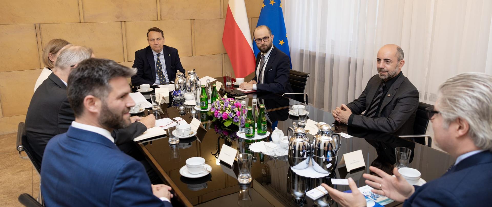 Spotkanie delegacji przy prostokątnym stole konferencyjnym. Na stole znajdują się dokumenty, mikrofony, filiżanki, szklanki, karafki z wodą oraz kompozycja z różowych i fioletowych kwiatów. W tle widoczne są flagi Polski i Unii Europejskiej.