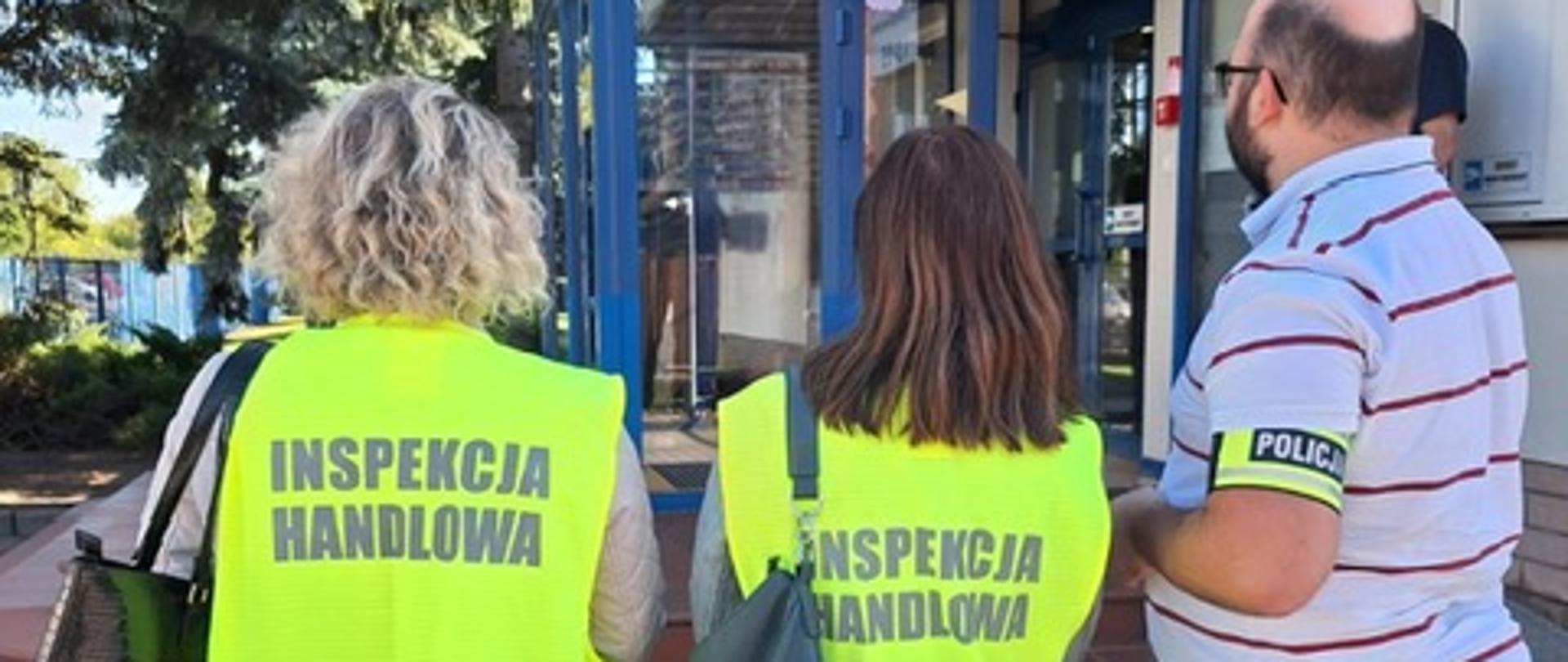 Dwie inspektorki Inspekcji Handlowej w kamizelkach odblaskowych w asyście policjanta stojące przed wejściem do budynku.