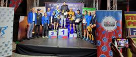 Zwycięzcy biegu stoją na podium obok organizatorzy biegu. Zwycięzcy prezentują nagrody. W tle ścianka okolicznościowa organizatora biegu. Po bokach zdjęcia roll-upy współorganizatorów. Uczestnicy stoją na scenie o konstrukcji rurowej. 
