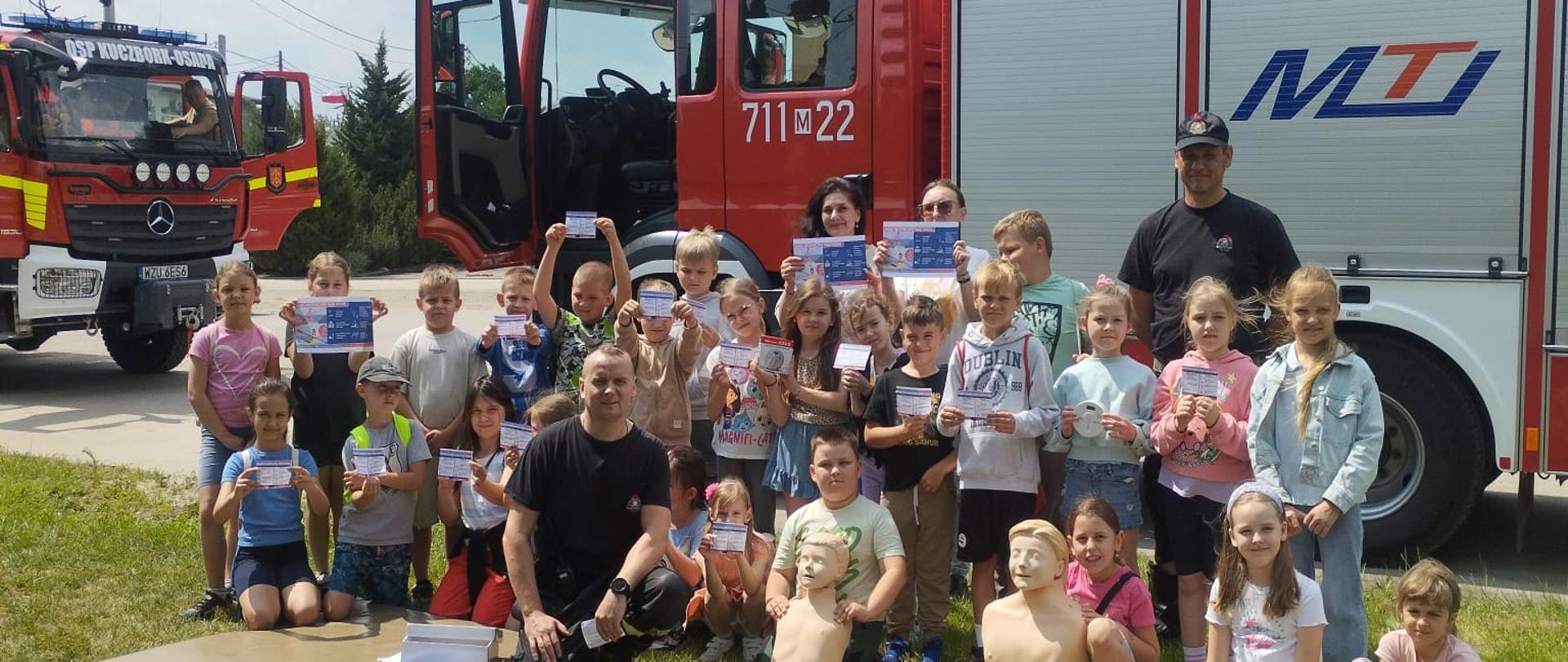 Na zdjęciu widoczna jest duża grupa dzieci, dorośli oraz dwa wozy strażackie. Scena rozgrywa się na świeżym powietrzu, na trawniku, z budynkami i drzewami w tle. W centrum zdjęcia znajduje się grupa dzieci. Obok nich, pośrodku i po prawej stronie, widać dwóch strażaków, ubranych w ciemne koszulki. Jeden z nich klęczy obok dzieci. Przed dziećmi leżą na ziemi dwa manekiny do resuscytacji (jeden górnej części ciała, drugi przypominający dziecko) oraz inne akcesoria, co wskazuje, że odbywał się pokaz lub szkolenie z pierwszej pomocy.