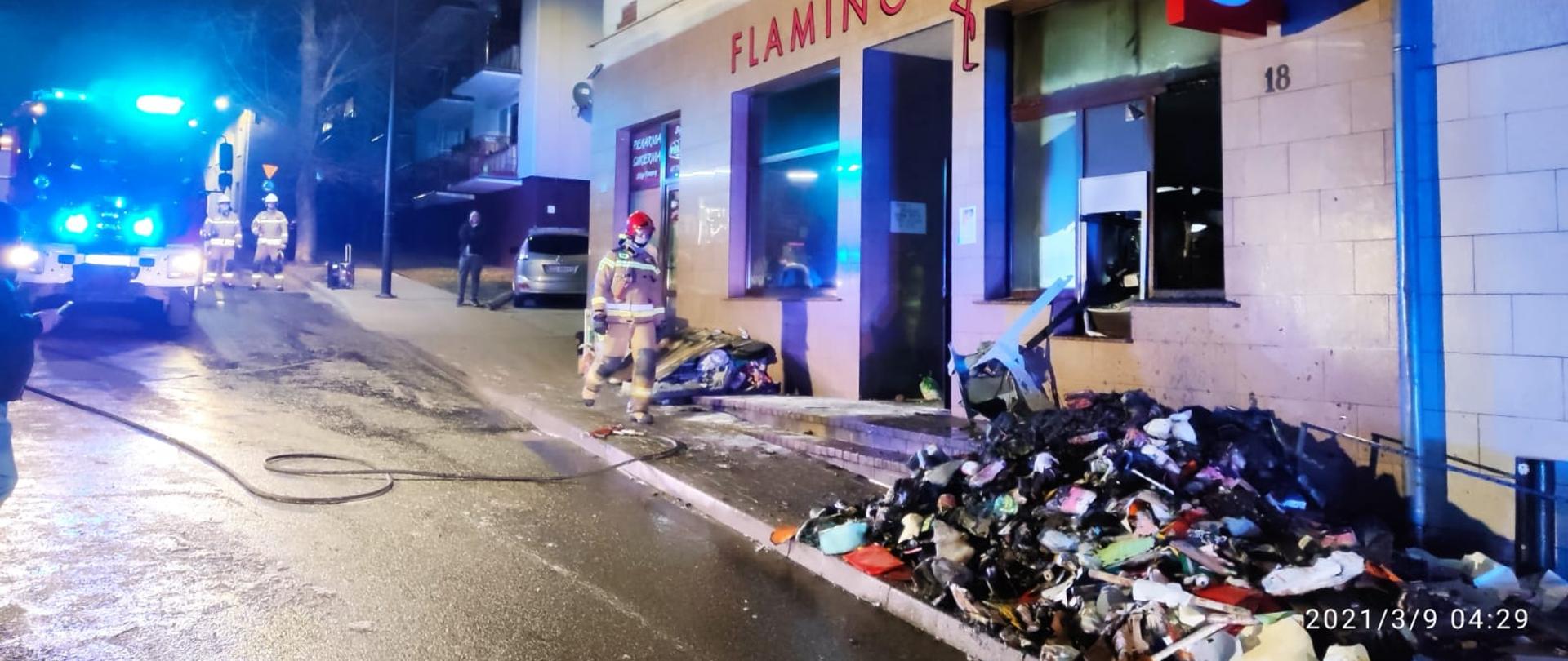 Na zdjęciu widoczny jest strażak, który idzie w kierunku sklepu, w którym doszło do pożaru, na ziemi rozwinięta linia gaśnicza. Na zewnątrz sklepu znajdują się nadpalone elementy wyniesione ze sklepu. W tle widać wozy strażackie oraz dwóch strażaków. 