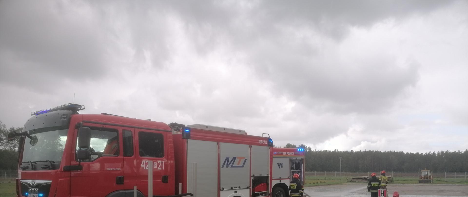 Pojazdy pożarnicze stoją na placu. Strażacy za pomocą linii wężowej podają prąd wody.