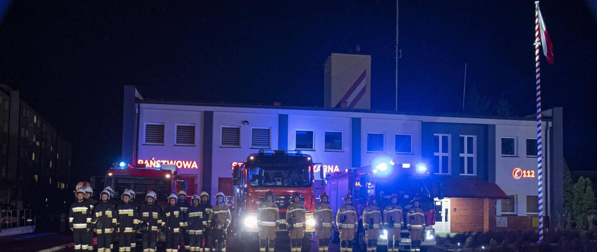 Punktualnie o godzinie 18 przed obiektem KP PSP zebrali się funkcjonariusze oraz druhowie z jednostki OSP by oddac hołd zmarłej koleżance i koledze. Druhowie z OSP Czernikowo zginęli w wypadku jadąc alarmowo do zdarzenia. Strażacy stoją w szyku przed autami pożarniczymi, które mają włączone sygnały świetlne i dzwiekowe. W tle budynek komendy powiatowej.