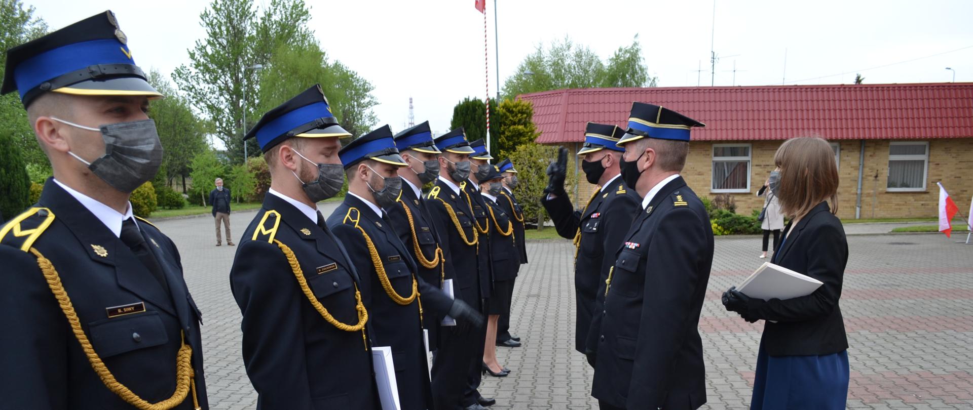 Strażacy stojący w rzędzie odbierają nagrody i wyróżnienia z rąk dwóch strażaków komendantów, jeden z nich salutuje, z boku kobieta cywil podająca nagrody. 