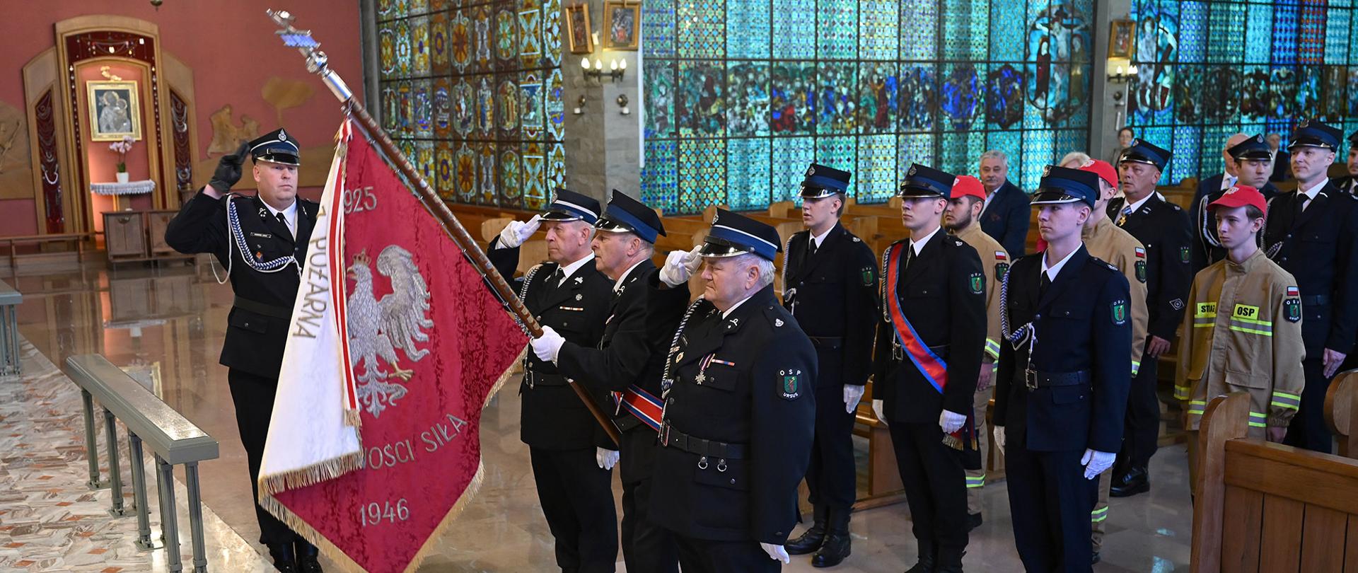 Strażacy z OSP Ursus ubrani w mundury wyjściowe. Z przodu stoi poczet sztandarowy. Wszyscy są zwróceni w stronę ołtarza, oddają honory. W tle widoczny jest ołtarz z obrazem i duże witraże, przez które wpada światło.