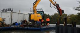 Na stawie, tuż przy brzegu wyspy, na specjalnej pływającej platformie znajduje się koparka i pracownicy firmy budowlanej. 