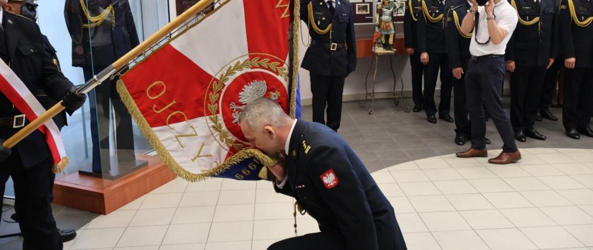 Zdjęcie wykonane w Sali tradycji komendy miejskiej Państwowej Straży Pożarnej podczas uroczystej zbiórki dotyczącej powołania na stanowisko komendanta miejskiego. Na zdjęciu widać moment pożegnania ze sztandarem komendanta miejskiego Państwowej Straży Pożarnej w Radomiu. Funkcjonariusz klęczy na jednym kolanie przed pochylonym sztandarem. Jedną ręką trzyma róg sztandaru składając na niego pocałunek. Pochylony sztandar w rękach trzyma jeden z trzech funkcjonariuszy tworzących poczet sztandarowy. W tle widać pozostałych funkcjonariuszy oraz pracowników cywilnych komendy miejskiej.