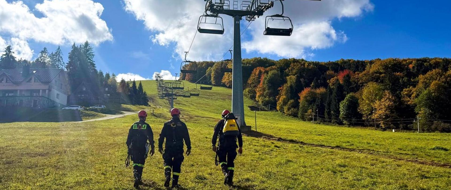 Zdjęcie przedstawia trzech strażaków którzy ze sprzętem wysokościowym idą w górę wyciągu narciarskiego.